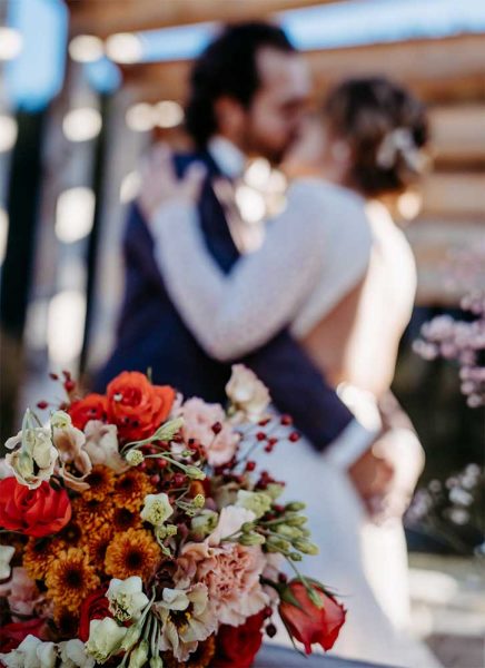 Ein roter Brautstrauß ist im Fokus. Im Hintergrund küsst sich ein Brautpaar.